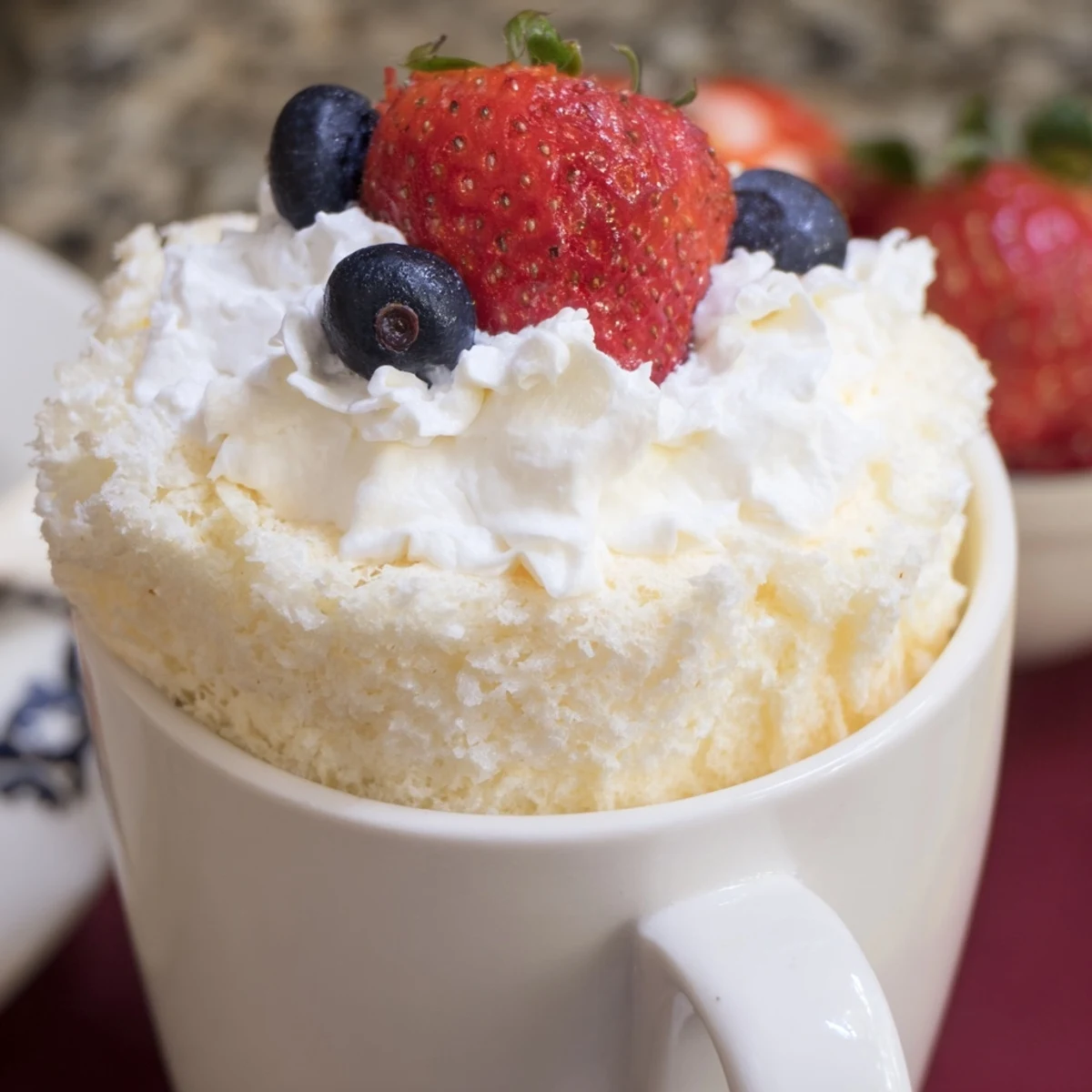 Golden fluffy angel food cake rises in a mug topped with dollop of white whipped cream and vibrant fresh red strawberries