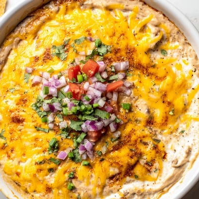Freshly baked Bean Dip with Cheddar Cheese, melted and gooey, served in a dish with crunchy tortilla chips and sliced jalapeños for dipping.